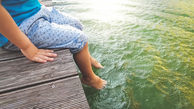 Nahaufnahmefoto des 3 Jahre alten kleinen Jungen, der auf dem hölzernen Pier sitzt und seine Füße im Flusswasser hält. Kind planschen im See mit Beinen