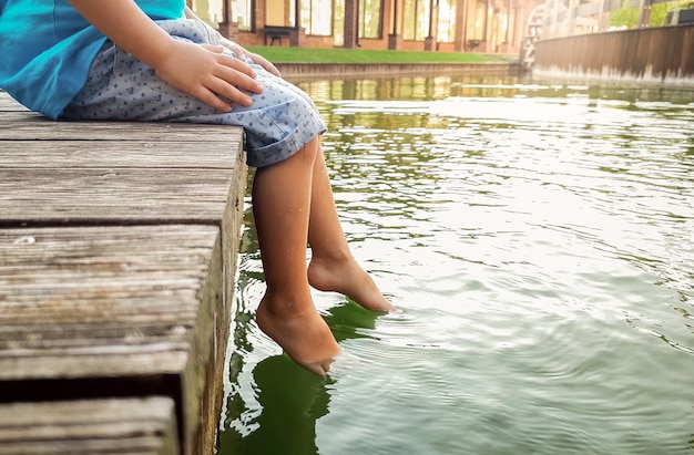 Nahaufnahmefoto des 3 Jahre alten kleinen Jungen, der auf dem hölzernen Pier sitzt und seine Füße im Flusswasser hält. Kind planschen im See mit Beinen