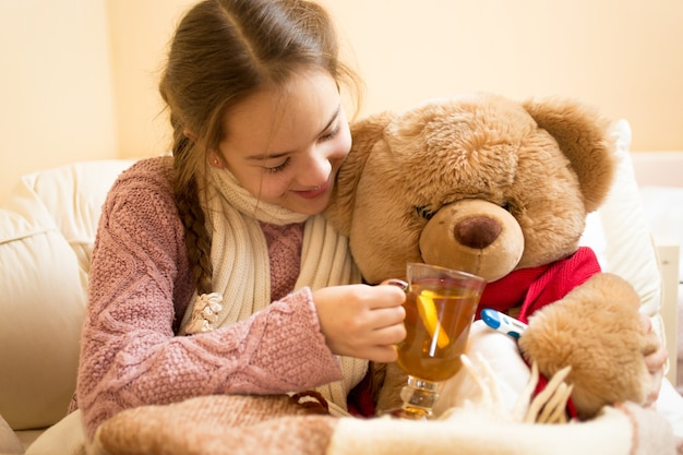 Nahaufnahmefoto des kleinen kranken Mädchens, das dem Teddybären heißen Tee gibt