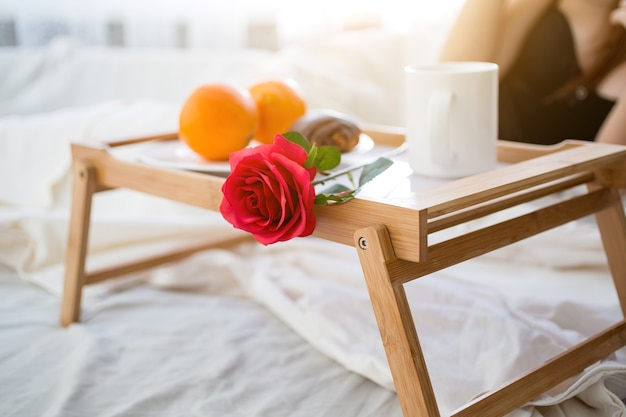 Nahaufnahmefoto des Tabletts mit Frühstück und roter Rose auf dem Bett im Hotelzimmer