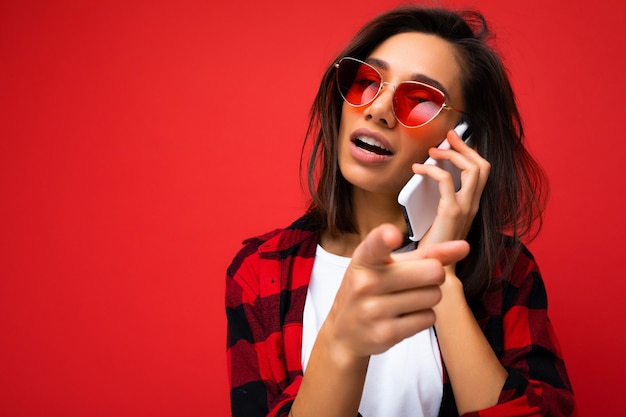 Nahaufnahmefoto einer attraktiven, positiven jungen Brunet-Frau, die ein stilvolles rotes Hemd, ein weißes T-Shirt und eine rote Sonnenbrille trägt, isoliert auf rotem Hintergrund, die auf dem Handy mit Blick auf die Seite und den Po kommunizieren