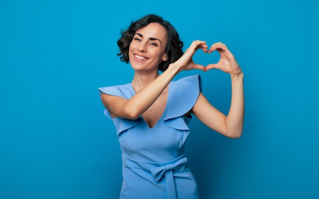 Nahaufnahmefoto einer fröhlichen süßen Frau in einem langen blauen Kleid, die in die Kamera schaut und lächelt, während sie ein Herz mit ihren Händen isoliert auf blauem Hintergrund zeigt