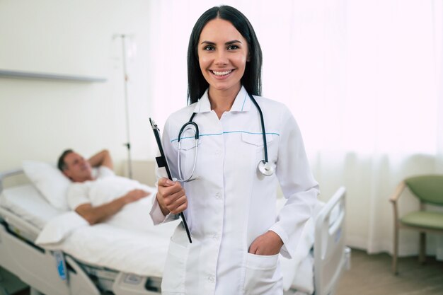 Nahaufnahmefoto einer lächelnden selbstbewussten niedlichen Ärztin in einem Mantel mit einem Stethoskop ist mit dem Patienten im Bett in der