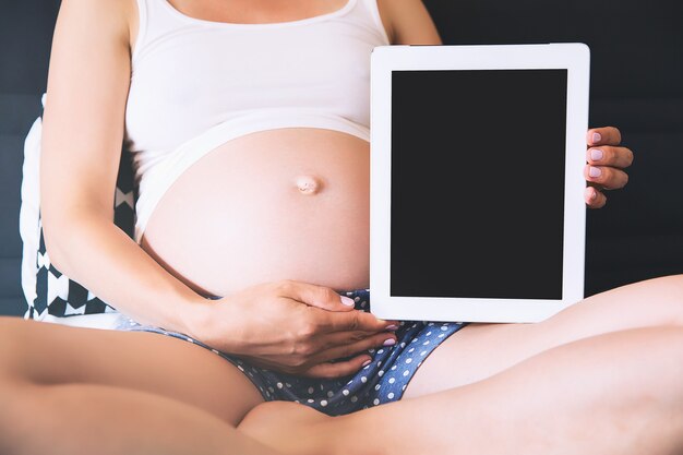 Nahaufnahmefoto einer schwangeren Frau, die zu Hause einen leeren Tablet-Bildschirm hält und zeigt