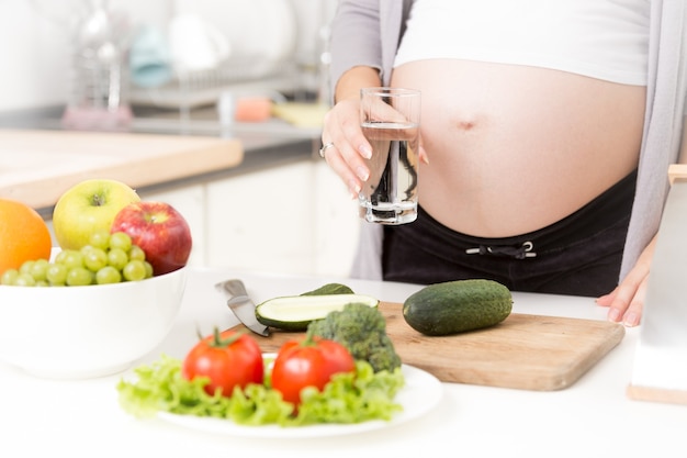 Nahaufnahmefoto einer schwangeren Frau mit einem Glas Wasser in der Küche