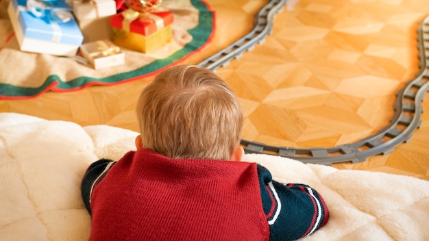 Nahaufnahmefoto eines kleinen Jungen, der auf dem Boden liegt und seine Spielzeugeisenbahn im Wohnzimmer auf Eisenbahnen betrachtet