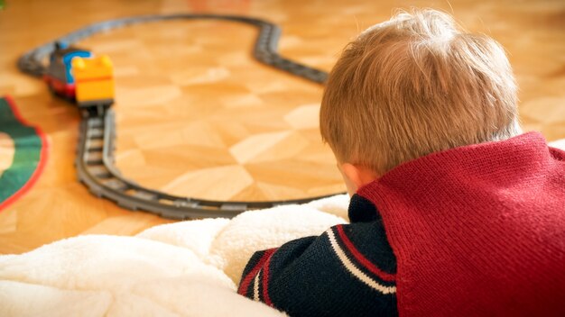 Nahaufnahmefoto eines kleinen Jungen, der seine Spielzeugeisenbahn auf Eisenbahnen auf Holzboden im Haus beobachtet