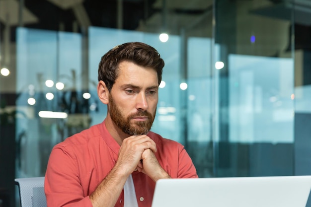 Nahaufnahmefoto konzentrierter und nachdenklicher junger Mann in einem roten Hemd arbeitet Studien auf einem Laptop im