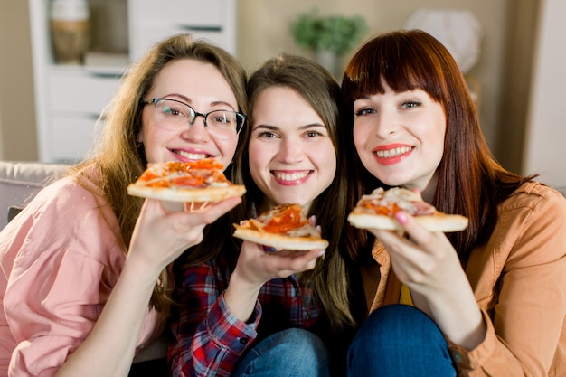 Nahaufnahmefoto von drei fröhlichen lächelnden Mädchen, die eine Party zu Hause feiern und Pizza essen.