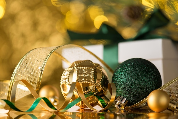 Nahaufnahmefoto von goldenen und grünen Christbaumschmuck-Ornamentenbällen, Serpentinenband, weißer Geschenkbox mit Schleife und Tannenzweig auf goldenem Bokeh-Hintergrund