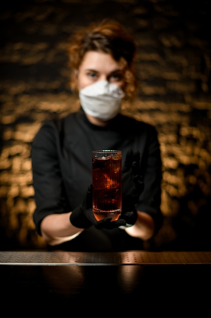 Nahaufnahmefrau an der Bar in der medizinischen Maske und in den schwarzen Handschuhen präsentieren Glas mit Cocktail