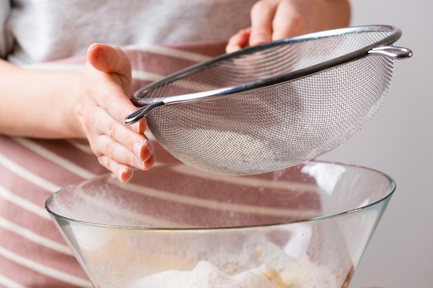 Nahaufnahmefrau siebt Weizenmehl durch ein Sieb in eine Rührschüssel für die Teigzubereitung für Kuchen und