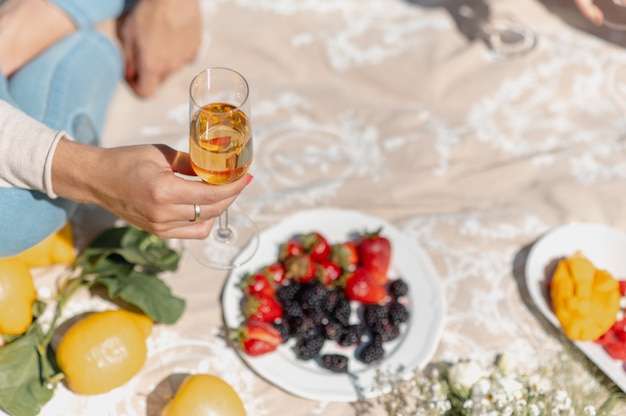 Nahaufnahmefrauenhand, die ein glas weißwein sitzt auf einem deckenpicknick mit früchten hält.