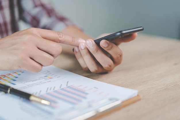 Nahaufnahmegeschäftsmann, der einen stift, ein smartphone hält und auf das finanzielle diagramm zeigt, das geschäftsbericht auf hölzernem schreibtisch mit computerlaptop neben zu hause überprüft. Stock Foto