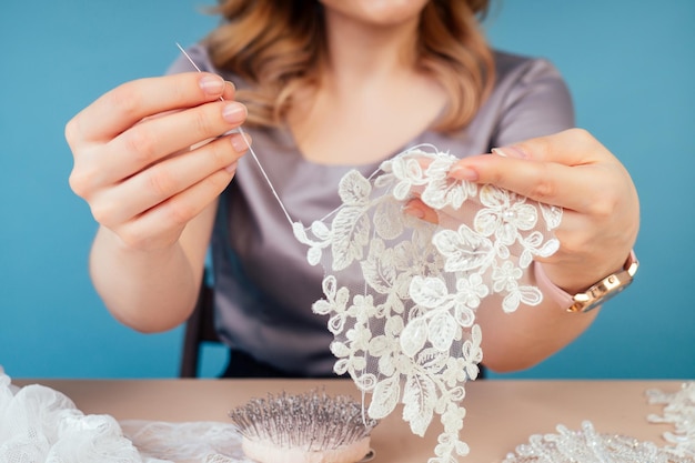 Nahaufnahmehände der Schneiderin Schneiderin Designer Hochzeitskleid näht Perlen zum Schnüren auf blauem Hintergrund im Studio.