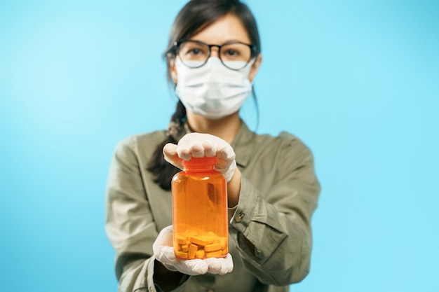 Nahaufnahmehände einer jungen Frau in einer medizinischen Schutzmaske und in den Handschuhen, die eine orange Dose mit Pillen auf einem blauen Hintergrund halten. Prävention oder Behandlung von Viren und Grippe