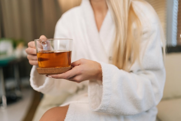 Nahaufnahmehände einer nicht erkennbaren blonden Frau, die einen weißen Bademantel trägt und eine Tasse mit frischem Kräutertee hält, die nach den Behandlungsverfahren auf dem Sofa im Spa-Salon sitzt