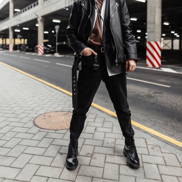 Nahaufnahmekörper eines jungen Mannes in einer übergroßen stilvollen Lederjacke mit einem Gürtel in einem klassischen Hemd in trendigen Jeans in coolen saisonalen Stiefeln in der Nähe einer Straße in der Stadt. Modischer Typ. Amerikanischer Stil.