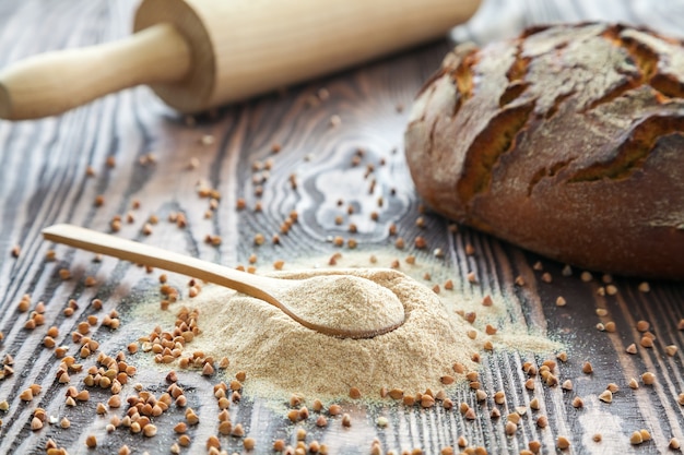 Nahaufnahmelöffel mit Buchweizenmehl, einem Brot und einem Nudelholz auf einem hölzernen Hintergrund. Alternatives Mehl. Glutenfreie und gesunde Ernährung.