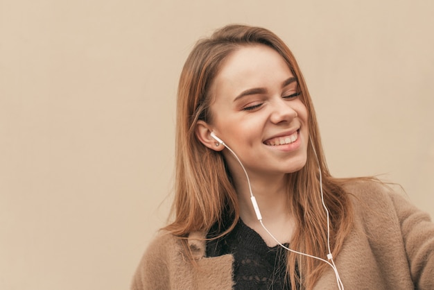 Nahaufnahmeporträt, das Musik in ihren Kopfhörern mit ihren geschlossenen Augen und lächelndem beigem Wandhintergrund hört