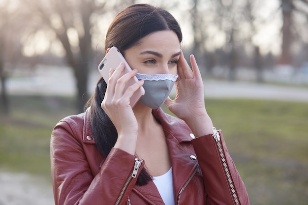 Nahaufnahmeporträt der besorgten Frau, die Schutzmaske und stilvolle Lederjacke trägt, die mit Telefon sprechen