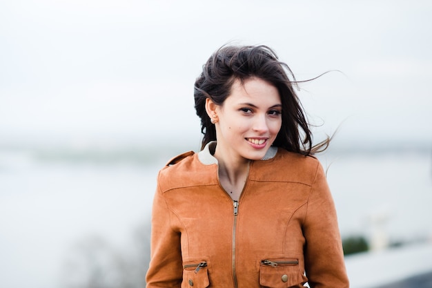 Nahaufnahmeporträt der jungen glücklichen schönen attraktiven Frau tragenden Ledermantel lächelnd und stehend über hellem Außenhintergrund. Wind weht ihr die Haare.