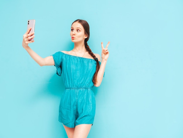 Nahaufnahmeporträt der jungen schönen lächelnden Frau im modischen Sommerjeansoverall Sexy sorglose Frau mit Schwanzfrisur, die nahe Wand im Studio aufwirft Positives Modell, das Spaß hat, selfie nehmend