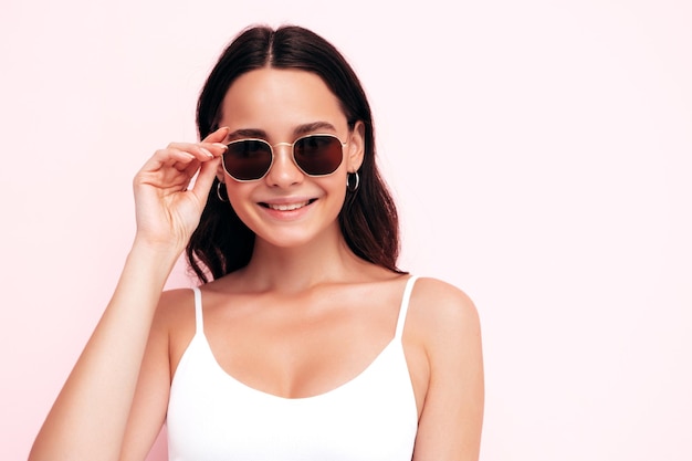 Nahaufnahmeporträt der jungen schönen lächelnden Frau in der modischen Sommerkleidung Sexy sorglose Frau, die nahe rosa Wand im Studio aufwirft Positives Modell, das Spaß drinnen hat Sonnenbrille ausziehen