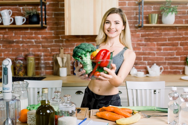 Nahaufnahmeporträt der lächelnden jungen Frau, die Glasschale mit frischem Gemüse beim Stehen am Tisch mit gesundem Essen in der modernen Küche hält. Gesundes Lebensmittelkonzept