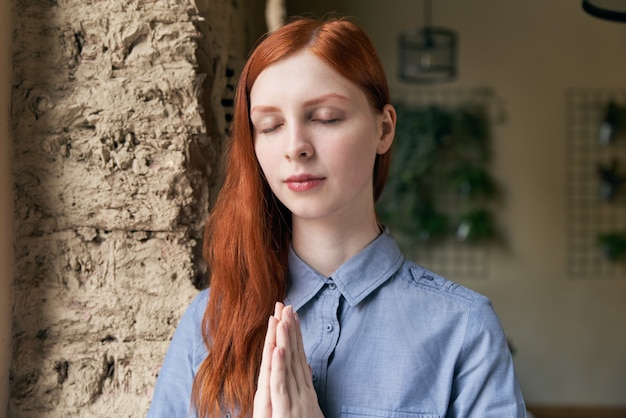 Nahaufnahmeporträt der langhaarigen rothaarigen Frau, die für ein Porträt mit geschlossenen Augen und gefalteten Händen vor dem Gesicht in der Haltung des Gebets oder der Meditation aufwirft