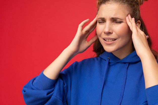 Nahaufnahmeporträt der müden müden jungen attraktiven blonden Frau mit zwei Hörnern mit aufrichtigen Gefühlen, die den stilvollen Hoodie einzeln über rotem Hintergrund mit Kopienraum tragen und Kopfschmerzen haben. Schmerzkonzept