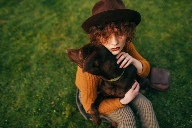 Nahaufnahmeporträt der stilvollen Dame im Hut und mit dem gelockten Haar, das Hund auf Draufsicht des Rasenhintergrundes umarmt