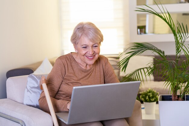 Nahaufnahmeporträt der zufälligen älteren Frau, die ihren Laptop beim Sitzen auf der Couch und beim Arbeiten verwendet. Attraktive Geschäftsfrau mittleren Alters, die vor Laptop sitzt und ihr kleines Geschäft von zu Hause aus verwaltet.