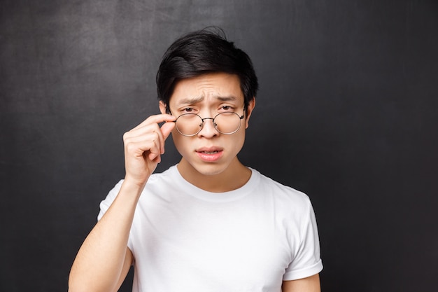 Nahaufnahmeporträt des arroganten skeptischen asiatischen Mannes im weißen T-Shirt, zusammengekniffen mit Zweifel abnehmende Brille, um Person näher zu betrachten, die seltsamen Unsinn spricht, stehend