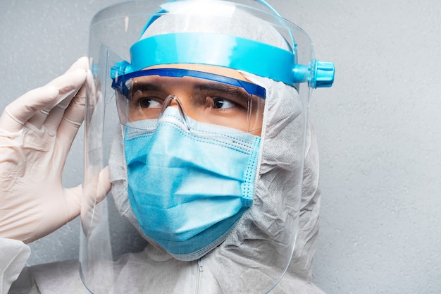 Nahaufnahmeporträt des jungen Doktormanns, der wegschaut, PSA-Anzug gegen Coronavirus und covid-19 tragend, auf Wand der grauen Wand. Pandemie-Konzept.