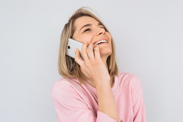 Nahaufnahmeporträt des positiven schönen blonden weiblichen Modells, das glücklich ist, mit Freund über Handy zu sprechen und zu lachen, das über weißer Wand aufwirftHübsche kaukasische Frau mit erfreutem Ausdruck mit Smartphone