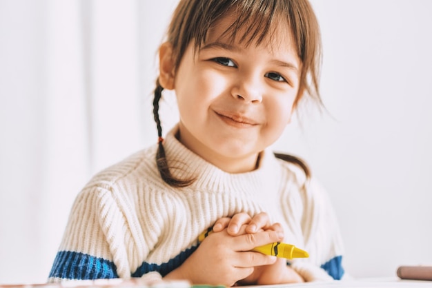 Nahaufnahmeporträt des schönen glücklichen kleinen Mädchens malt mit Ölbleistiften, die zu Hause am weißen Schreibtisch sitzen Hübsches lächelndes Vorschulkind, das mit gelbem Bleistift aufwirft Menschen-Kindheits-Bildungskonzept