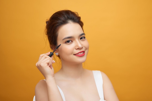 Nahaufnahmeporträt einer attraktiven jungen Frau, die etwas Mascara auf ihre Wimpern legt, mit Make-up-Pinsel über gelbem Hintergrund