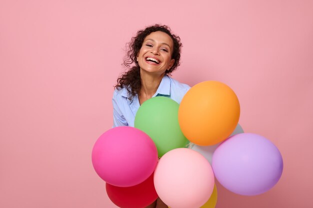 Nahaufnahmeporträt einer charmanten fröhlichen gemischtrassigen hispanischen Frau mit bunten bunten Luftballons, lachend in die Kamera schauend, einzeln auf rosafarbenem Hintergrund mit Kopienraum.