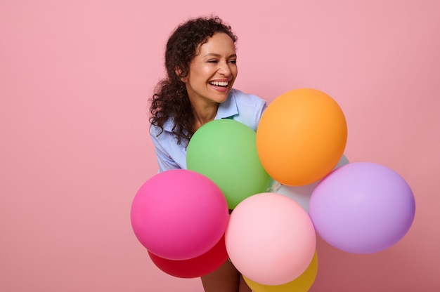 Nahaufnahmeporträt einer entzückenden, fröhlichen afroamerikanischen Frau mit gemischten Rassen mit bunten bunten Ballons, die zur Seite lachen, einzeln auf rosafarbenem Hintergrund mit Kopienraum.