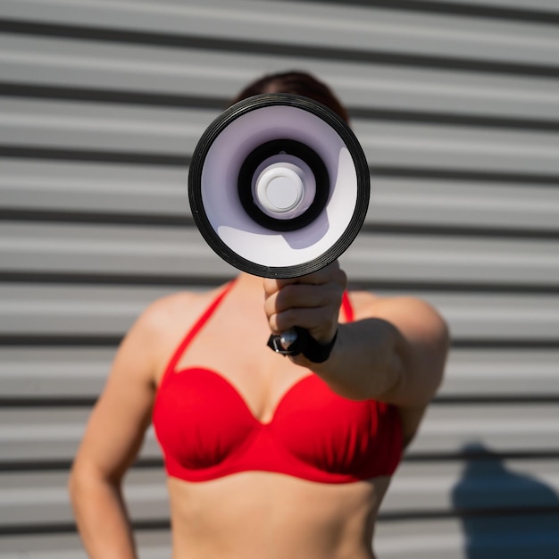 Nahaufnahmeporträt einer Frau in einem roten Bikini, die ein Megaphon vor dem Hintergrund ihres Gesichts hält. Ein Mädchen in einem Badeanzug macht eine Durchsage über den Lautsprecher vor dem Hintergrund eines Stahlblechs