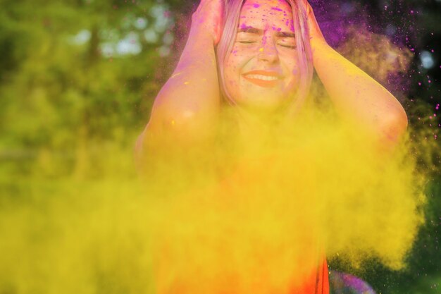 Nahaufnahmeporträt einer fröhlichen blonden Frau, die Spaß in einer Wolke aus gelber und lila trockener Holi-Farbe hat