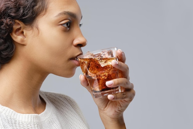 Nahaufnahmeporträt einer jungen afroamerikanischen Frau, die Cola mit Eis auf grauem Hintergrund trinkt