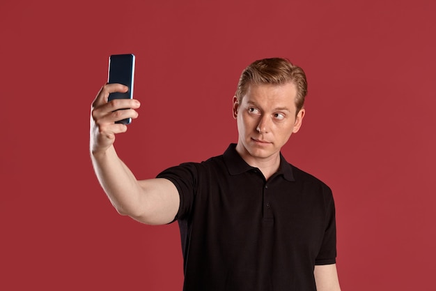 Nahaufnahmeporträt einer jungen, eleganten rothaarigen Person in einem stilvollen schwarzen T-Shirt, die ein Selfie macht, während sie auf rosafarbenem Studiohintergrund posiert. Menschliche Gesichtsausdrücke. Aufrichtiges Gefühlskonzept. Platz kopieren.