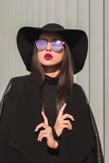 Nahaufnahmeporträt einer luxuriösen brünetten Frau in modischem Mantel, trägt Hut und Brille und posiert im Sonnenlicht