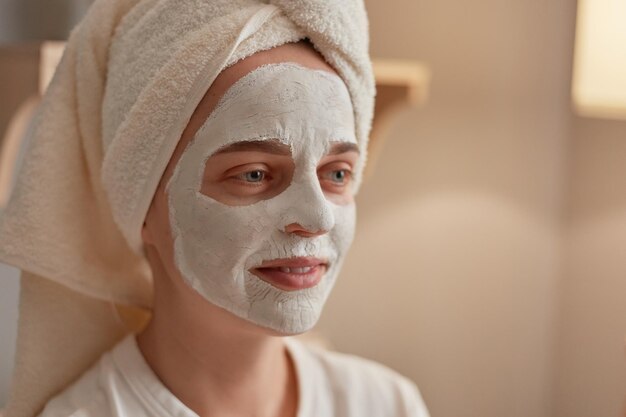 Nahaufnahmeporträt einer schönen Frau mit Gesichtsmaske aus weißem Ton auf dem Gesicht, die in ein Handtuch gewickelt wird und am Wochenende zu Hause Hautpflege- und Schönheitsbehandlungen durchführt