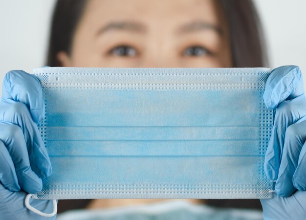 Nahaufnahmeporträt einer schönen jungen asiatischen Frau, die ihr Gesicht mit einer medizinischen blauen Gesichtsmaske bedeckt