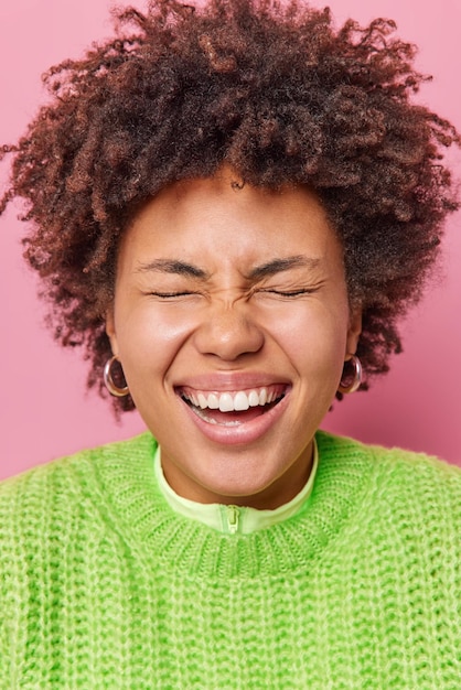 Nahaufnahmeporträt einer überglücklichen jungen Frau, die die Augen geschlossen hält, kichert positiv lächelt und zeigt weiße Zähne trägt einen warmen grünen Strickpullover einzeln auf rosafarbenem Hintergrund. Aufrichtige Gefühle