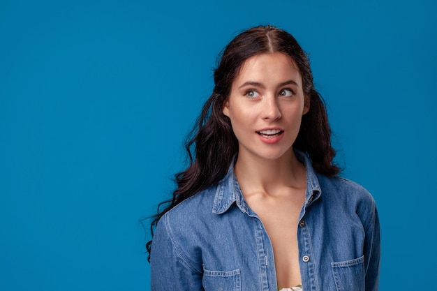 Nahaufnahmeporträt eines attraktiven Brunettemädchens mit dem langen gelockten Haar, das auf einem blauen Hintergrund aufwirft.