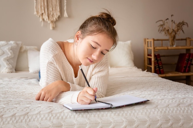 Nahaufnahmeporträt eines entspannten jungen Mädchens, das Buch schreibt, während es auf dem Bett liegt Foto in hoher Qualität